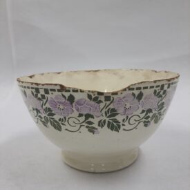 Tigela com exterior decorado com faixa de flores roxas claras estilizadas e folhagem verde escura sobre um fundo creme. Acima dessa faixa floral, há uma borda geométrica verde escura. A tigela está desgastada e danificada. A borda superior está irregular e com perdas em diversas partes, e uma superfície considerável foi deteriorada, revelando a cerâmica por baixo. O interior e o pé da tigela também apresentam descoloração e manchas.